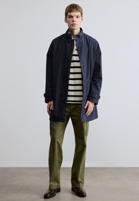 Meresinine veekindel mantel klassikalise krae, eessete nööpidega ja küljetaskutega, ühildatud triibulise kontrastse särgi ja oliivroheliste cargo-pükstega.