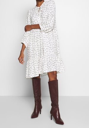 Kobieta ubrana w białą sukienkę do kolan z czarnym nadrukiem zebry i brązowe wysokie kozaki za kolano, stojąca na jednolitym tle.