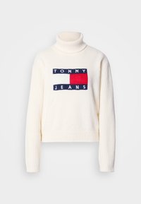 Pull à col roulé crème avec texture côtelée, arborant un logo "TOMMY JEANS" marine et rouge ainsi qu'un motif carré sur le devant.