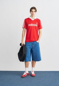 Κόκκινη φανέλα Adidas με λευκό γιακά και λογότυπο, σε συνδυασμό με μπλε τζιν σορτς. Το μοντέλο κρατάει ένα μαύρο μπουφάν, φοράει κόκκινα αθλητικά παπούτσια και λευκές κάλτσες.