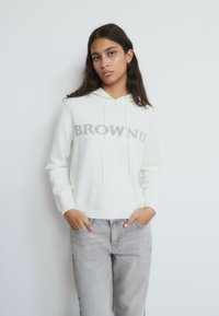 Sudadera blanca con texto "BROWNIE" en gris en el frente. Cuenta con capucha con cordón, mangas largas y un ajuste relajado, combinada con jeans gris claro.