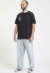 Sort bomulds-T-shirt med grafisk design på venstre bryst, kombineret med lyse, løstsiddende jeans og sorte sneakers.