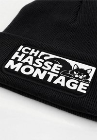 Black knitted beanie with a woven patch featuring a black cat illustration and the text "ICH HASSE MONTAGE" in bold white letters.