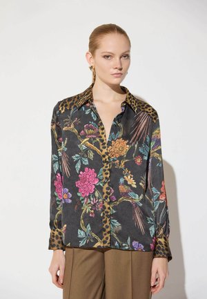 Mujer que lleva una blusa de manga larga con estampado floral y de pájaros, cuello y puños con estampado de leopardo, combinada con pantalones marrones de pierna ancha.