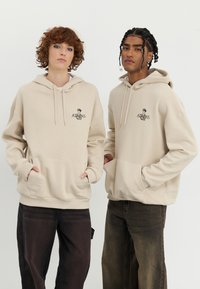 Beige hoodies with front pockets, featuring a black "ATHENS" logo. Model wears dark loose-fitting jeans. Soft fabric and relaxed design.