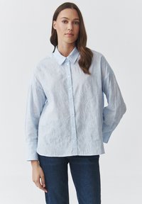 Camisa de botón de color azul claro hecha de tela texturizada, con bordado floral y un cuello clásico, combinada con jeans oscuros.