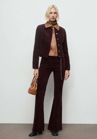 Chaqueta de pana burdeos con cuello marrón, combinada con pantalones acampanados a juego. Bolso marrón minimalista y botas negras hasta el tobillo.