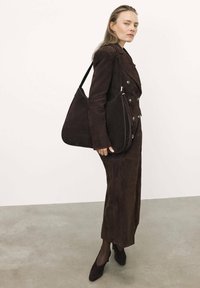Borsa a spalla in suede marrone dalla forma arrotondata, con piping a contrasto e una sola tracolla, indossata da un modello in un completo marrone abbinato.