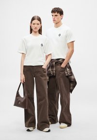 Fehér, kerek nyakú pólók átlós mintával a mellkason, barna bőszárú nadrággal és barna kézitáskával párosítva. Texturált cipők egészítik ki a megjelenést.