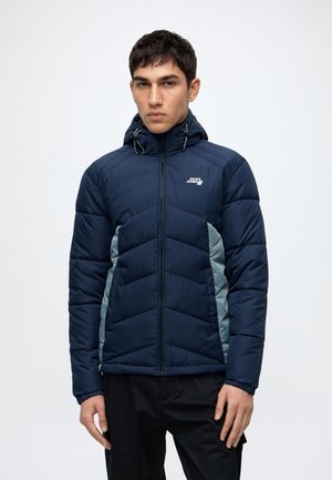 Homme portant une veste matelassée bleu marine avec une capuche et des panneaux latéraux gris clair, debout devant un fond uni.
