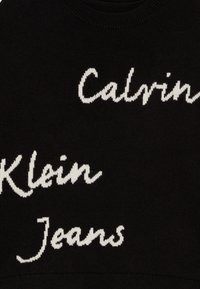 Schwarzer Strickpullover mit weißen Buchstaben. Die Worte "Calvin Klein Jeans" sind prominent in einer geschwungenen Schriftart dargestellt.