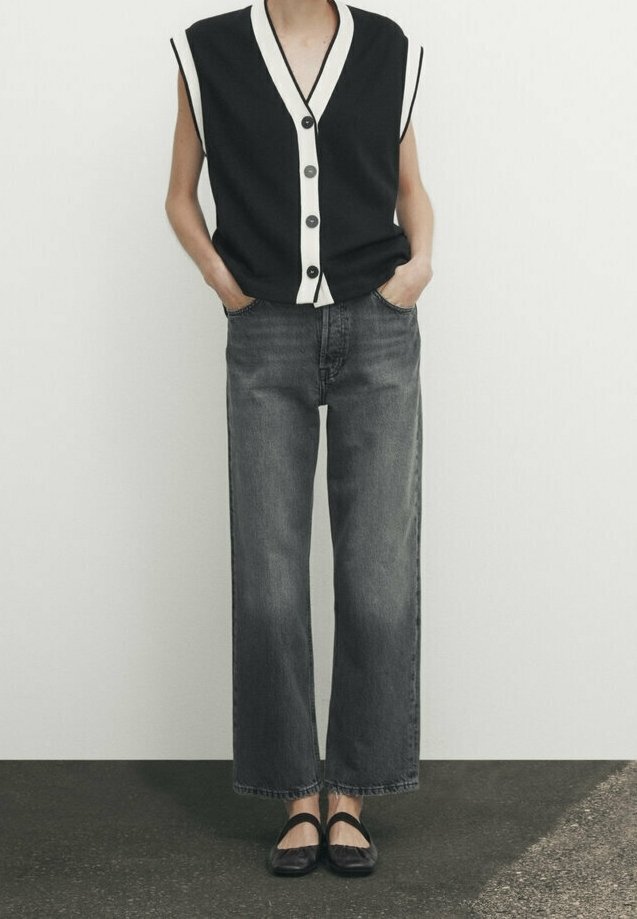 Personne portant un gilet noir et blanc sans manches boutonné, un jean noir délavé à jambes larges, et des chaussures Mary Jane noires, debout les mains dans les poches.