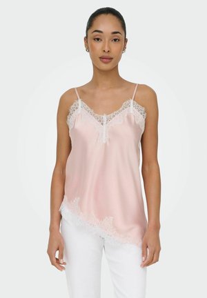 Donna che indossa una canottiera di raso rosa chiaro con bordo in pizzo bianco e pantaloni bianchi, in piedi davanti a uno sfondo chiaro semplice.