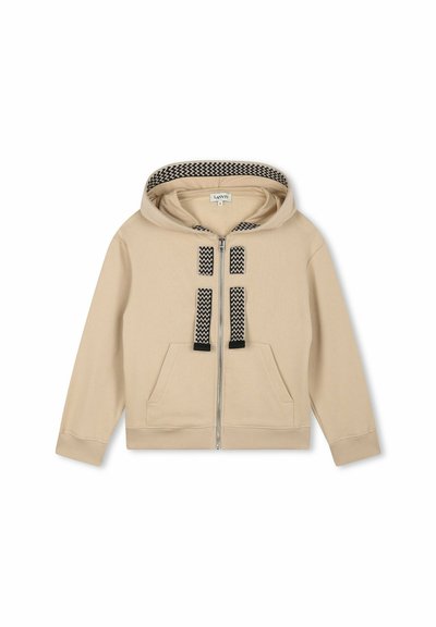 Haut à capuche beige zippé avec une doublure de capuche à motifs noir et blanc, deux poches avant et des sangles décoratives noires sur la poitrine.