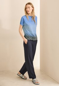 T-shirt bleu dégradé avec un motif texturé, associé à un pantalon bleu marine. Les sandales sont argentées avec des accents de boucles. Le modèle pose les mains dans les poches.