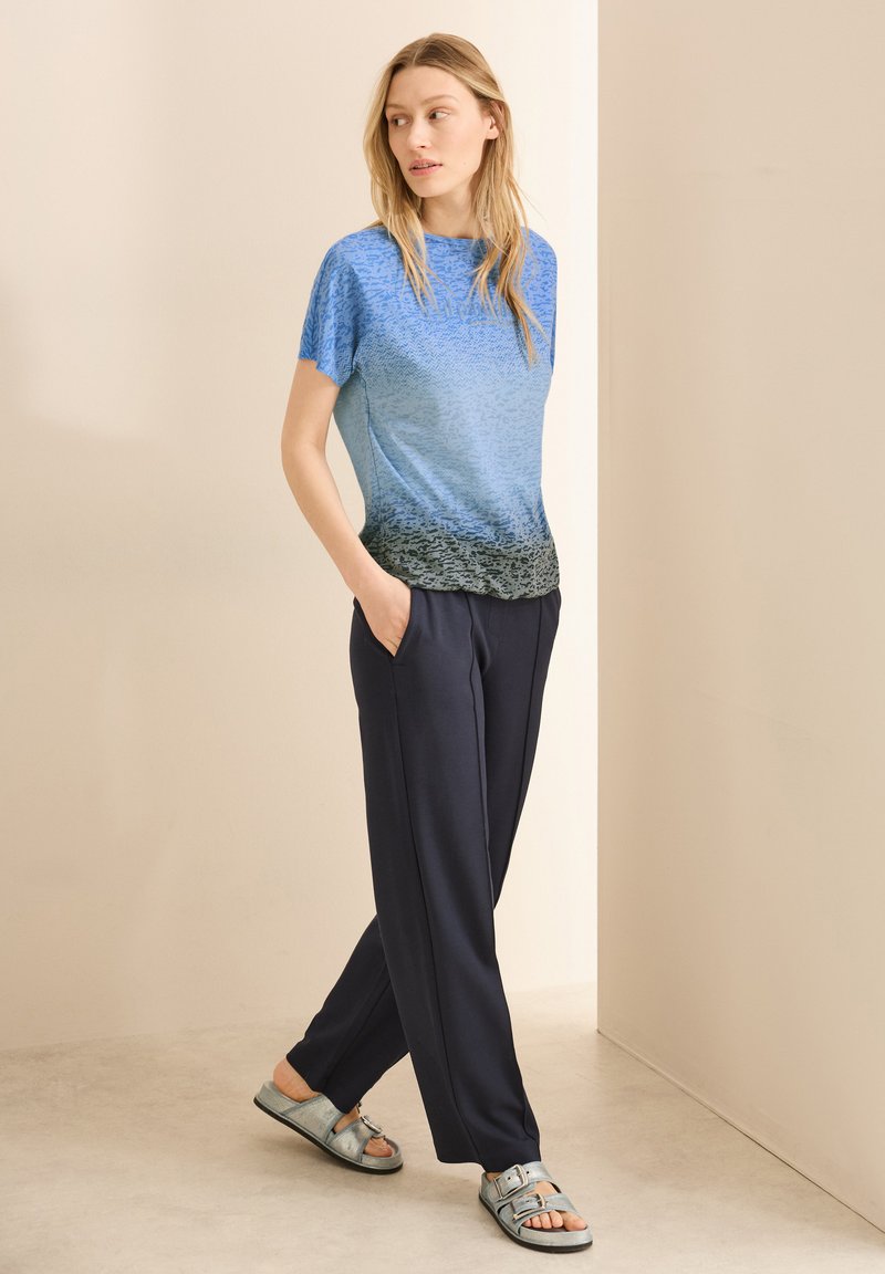 T-shirt bleu dégradé avec un motif texturé, associé à un pantalon bleu marine. Les sandales sont argentées avec des accents de boucles. Le modèle pose les mains dans les poches.