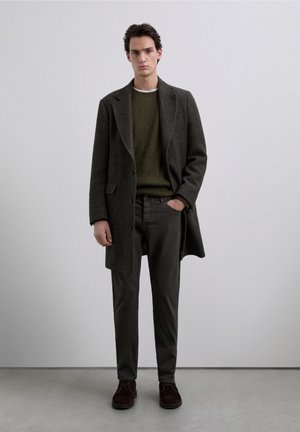 Abrigo largo de lana oscuro con solapas de punta sobre un suéter de punto acanalado verde oliva. Combinado con pantalones oscuros y zapatos de ante marrón.