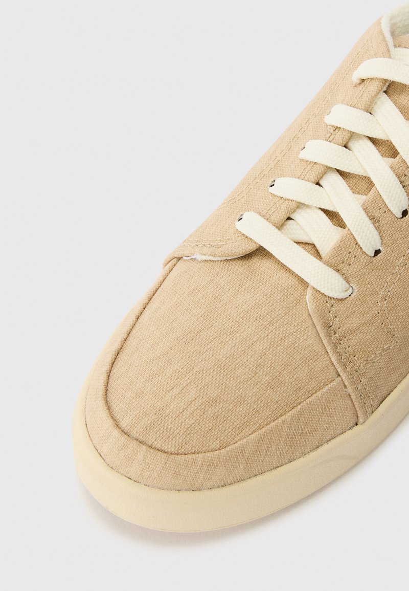 Sneaker in tela beige con lacci crema e punta rotonda. Il materiale presenta una superficie testurizzata con cuciture rinforzate lungo i bordi.