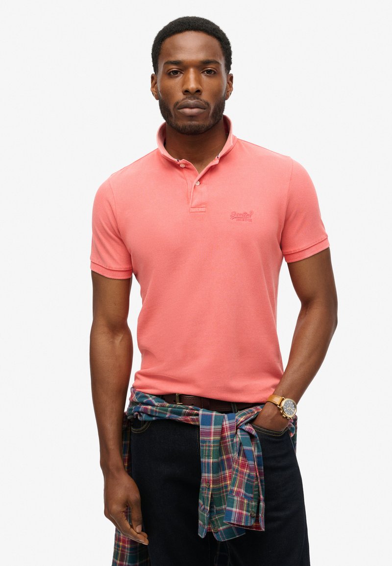 Camisa polo rosa con cuello, mangas cortas y logo bordado. Llevada sobre jeans oscuros con una camisa de cuadros atada a la cintura.