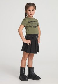 T-shirt verde oliva a maniche corte con testo grafico nero, abbinato a una gonna nera plissettata e stivaletti neri con cerniera e suole spesse.