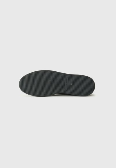 Semelle de chaussure en caoutchouc noir avec une surface texturée et des rainures horizontales, présentant un détail de logo rectangulaire près du talon.