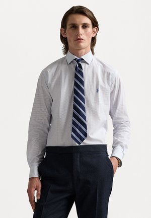 White shirt with a subtle pattern, paired with a dark striped tie. Slim fit, long sleeves, and a logo accent on the chest. Dark trousers.