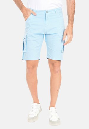 Uomo con pantaloncini cargo azzurri chiari e scarpe da ginnastica bianche, in piedi con una mano in tasca contro uno sfondo neutro.