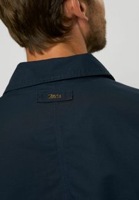 Chaqueta azul marino con una textura suave, que presenta un cuello clásico y una etiqueta bordada en dorado. Detalle de costura a lo largo del hombro.