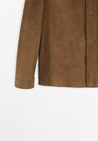 Détail d'une veste en daim marron à manches longues montrant les coutures et la fermeture par boutons sur un fond clair uni.