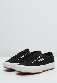 Black canvas sneakers with white rubber soles, featuring a red and white logo. Positioned on a light grey surface against a plain background.