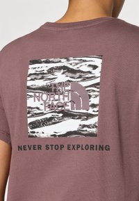 Bordeaux katoenen t-shirt met een grafisch ontwerp van bergen en het "THE NORTH FACE" logo in lichtpaars, voorzien van "NEVER STOP EXPLORING" in vetgedrukte zwarte tekst.