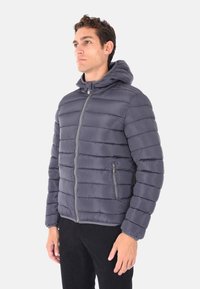 Uomo che indossa una giacca trapuntata grigio scuro con cappuccio e zip e pantaloni neri, in piedi contro uno sfondo chiaro e semplice.