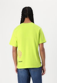 Neon grønn bomulls t-skjorte med korte ermer, med et logo-graphic i rosa på nedre rygg og svart tekst ved kanten.