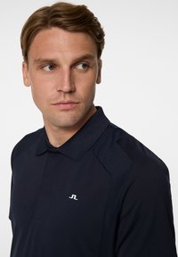 Marinblå pikétröja med spetskrage och korta ärmar. Har en liten vit logotyp på bröstet och texturerade axelpartier.