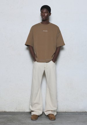 Un homme se tient les mains dans les poches, portant un t-shirt marron oversize, un pantalon large crème et des chaussures marron, devant un mur uni.