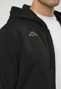 Melns rāvējslēdzēja džemperis ar kapuci, ar aukliņu, gludu tekstūru un baltu izšūtu logo krūtīs. Valkāts pār baltu kreklu.