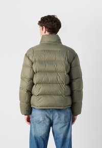 En ung person i en puffy olivengrønn jakke står med ryggen til kameraet, iført lyseblå jeans mot en ensfarget bakgrunn.
