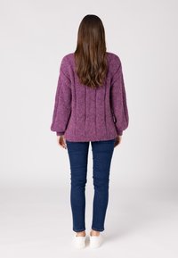 Lila gestrickter Pullover mit strukturiertem, geripptem Design; kombiniert mit blauen engen Jeans und weißen Sneakers. Sichtbares Haar reicht über die Schultern.