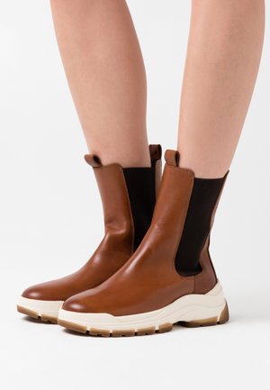 Stivaletti marroni in pelle con pannelli laterali elastici neri e suole in gomma beige. Presentano una punta arrotondata e una texture sottile.