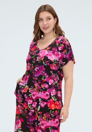 Vrouw draagt een zwart topje met korte mouwen en een broek met een felroze, rode en paarse bloemenprint, staand met één hand in de zak.