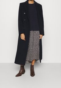Granatowy długi płaszcz z podwójnymi guzikami, w połączeniu z ciemnym swetrem dzianinowym i spódnicą w kwiatowy wzór. Brązowe botki za kostkę z teksturą krokodylej skóry.