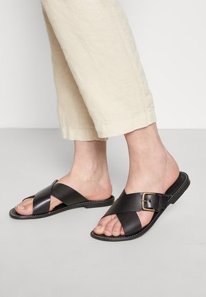 Schwarze Leder-Sandalen mit einem überkreuzten Riemendesign und einer Metallschnalle, flacher Sohle und dekorativen weißen Nähten am Rand.