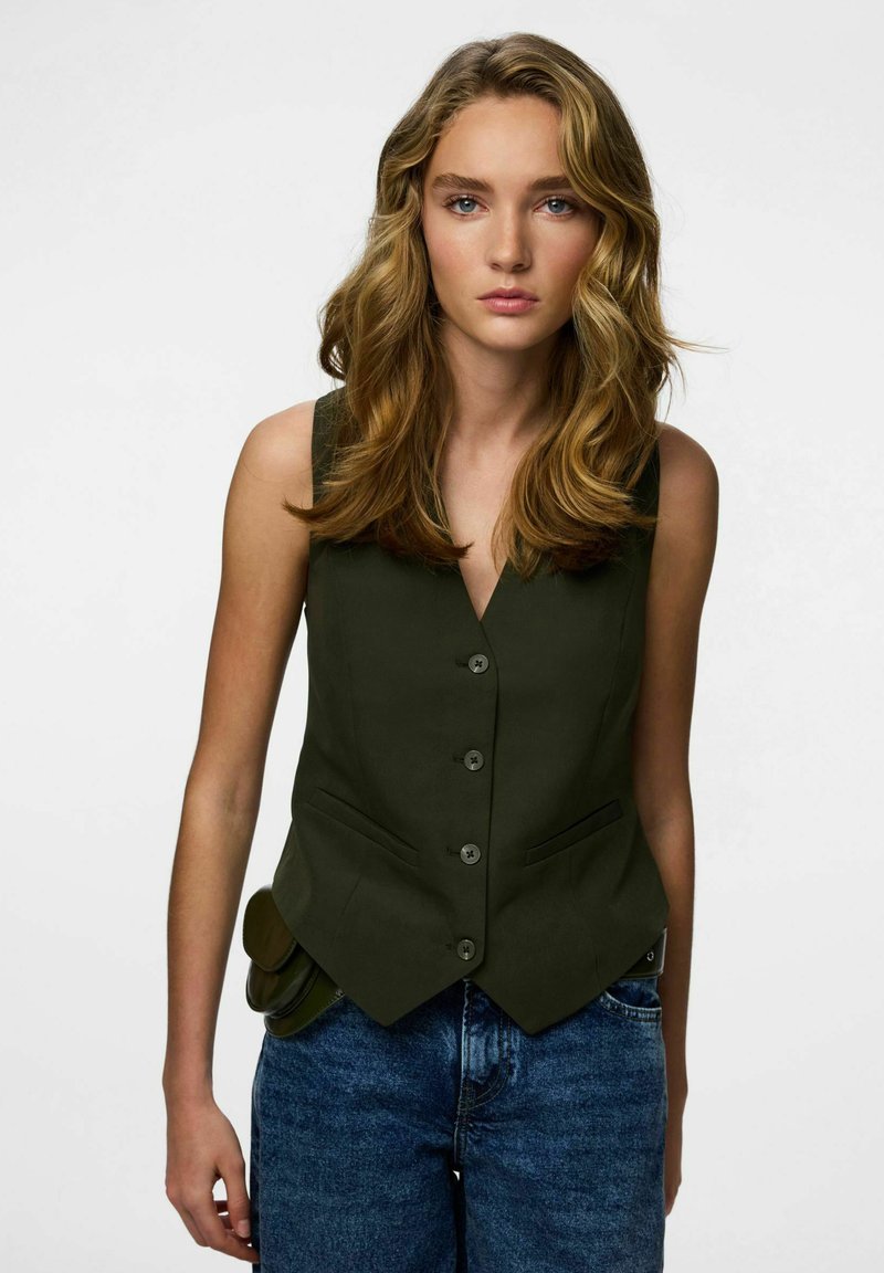 Tamnozeleni prsluk s V-izrezom, pet dugmadi i bočnim džepovima, uparen s plavim trapericama. Ima pripijeni kroj i glatku teksturu.