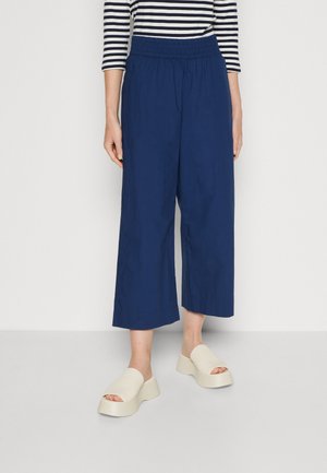 Personne portant un pantalon large court bleu marine, des sandales plates couleur crème à plateforme, et un haut à manches longues rayé noir et blanc, debout sur un sol clair.