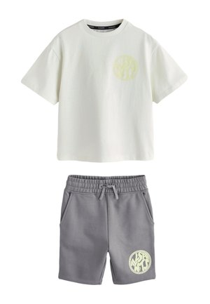 Junge mit kurzen Haaren, der ein übergroßes weißes T-Shirt, graue Shorts mit einem Logo, weiße Socken und weiße Turnschuhe trägt und vor einem einfarbigen Hintergrund steht.