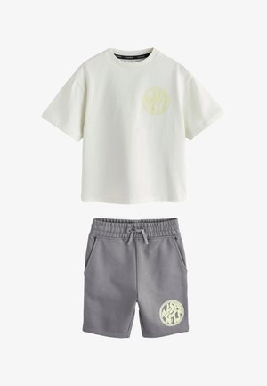 Junge mit kurzen Haaren, der ein übergroßes weißes T-Shirt, graue Shorts mit einem Logo, weiße Socken und weiße Turnschuhe trägt und vor einem einfarbigen Hintergrund steht.