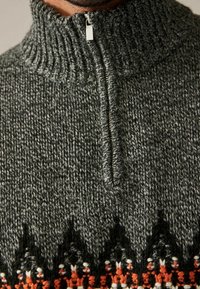 Close-up van een grijze gebreide trui met een hoge geribbelde kraag, zilveren rits en een zwart-orange patroon aan de onderkant.