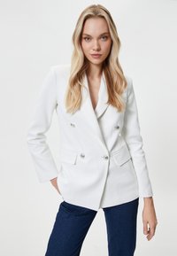 Blazer bianco doppiopetto in tessuto testurizzato, con bottoni argentati e due tasche frontali, abbinato a jeans blu scuro.