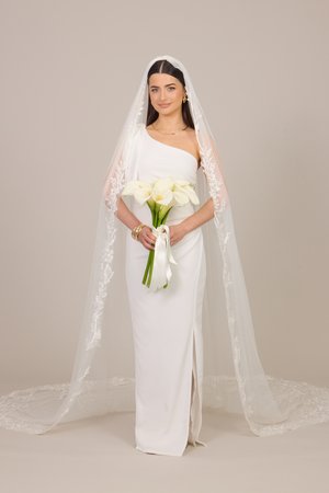 Ühe õlaga valges pulmakleidis naine pitsilise looriga hoiab käes valgete kalla-liiliate kimbu neutraalsel taustal.