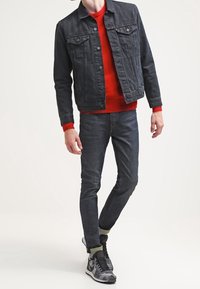 Czarna dżinsowa kurtka na czerwonym swetrze, w połączeniu z ciemnymi dżinsami i szarymi sneakersami z zielonymi akcentami. Model pozował w zrelaksowanej postawie.